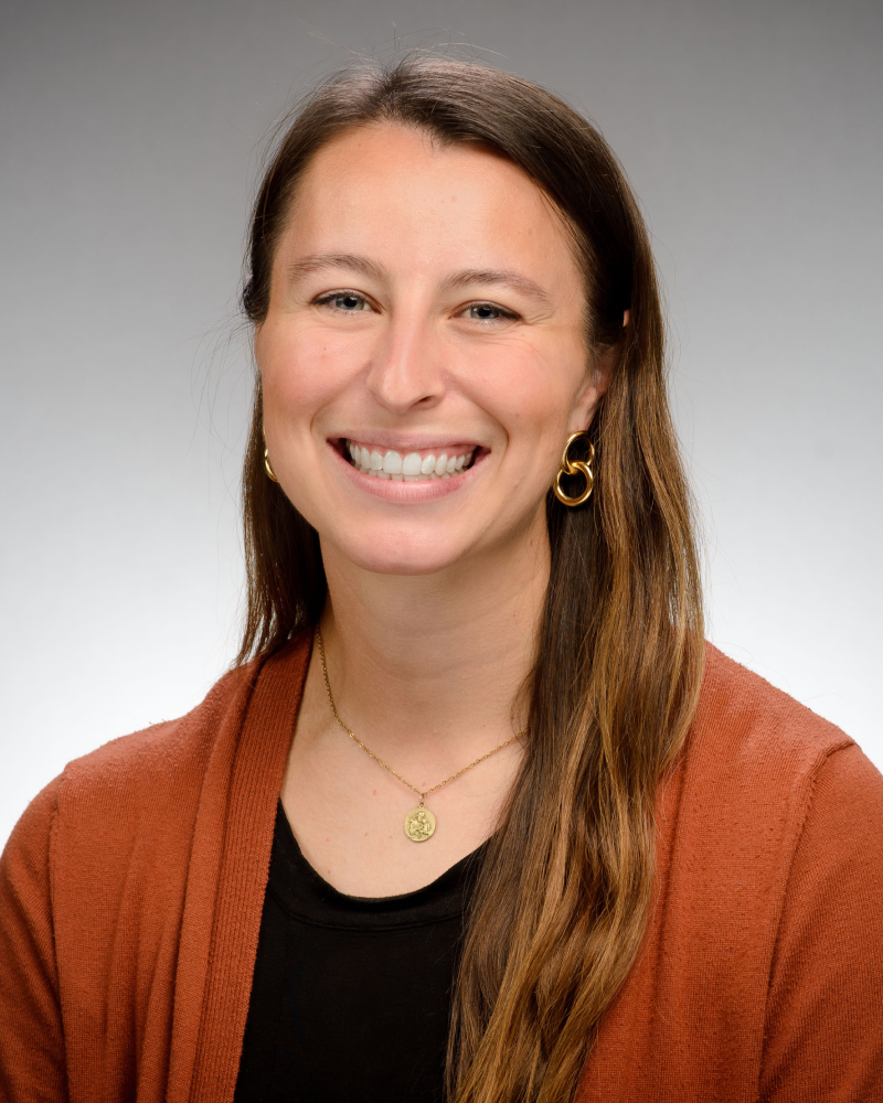 Headshot of Julia French, a woman with long brown hair, smiling, wearing an orange cardigan over a black top and a gold necklace.