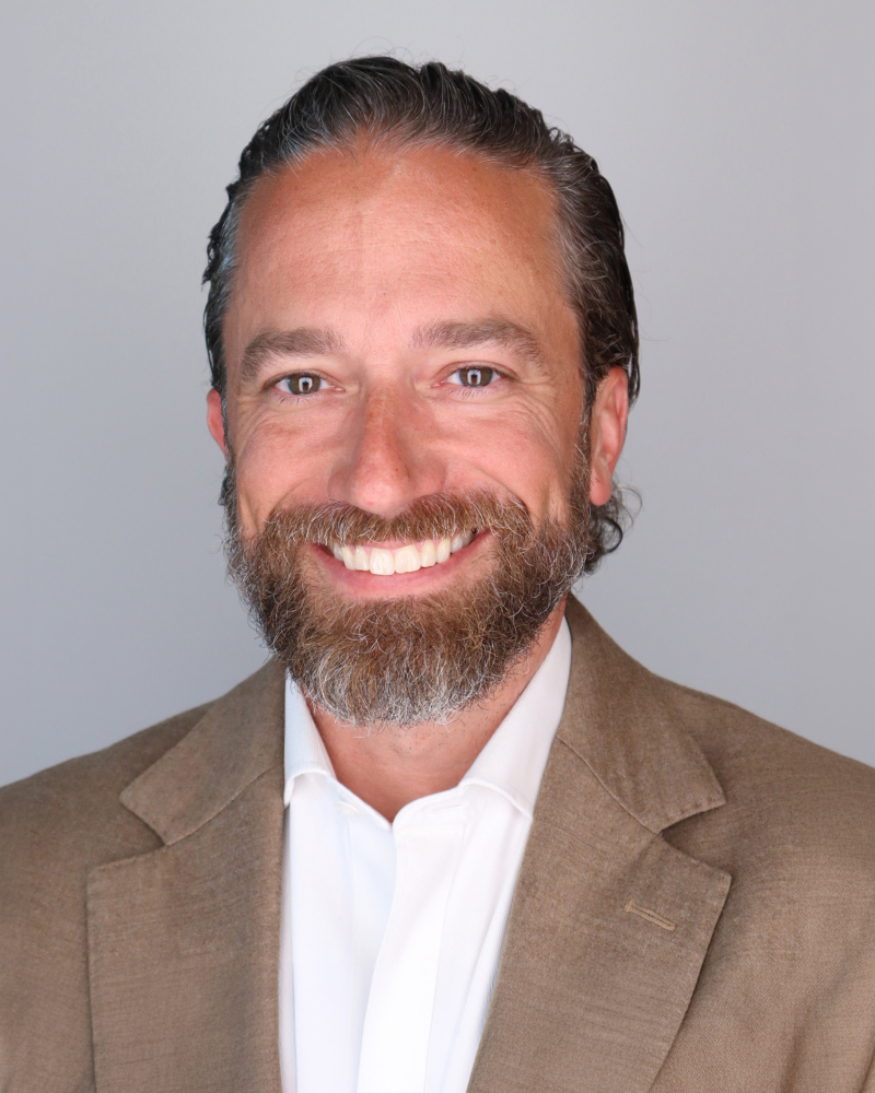 Headshot of Adam Kronk, a man with short dark hair and a gray beard, smiling, wearing a tan blazer over a white shirt.