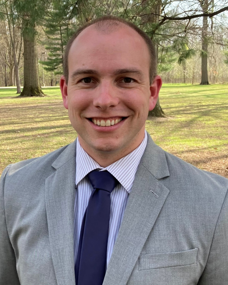 Headshot of Patrick Schmadeke, a man with short light hair, smiling, wearing a gray blazer, striped shirt, and navy tie, with trees in the background.