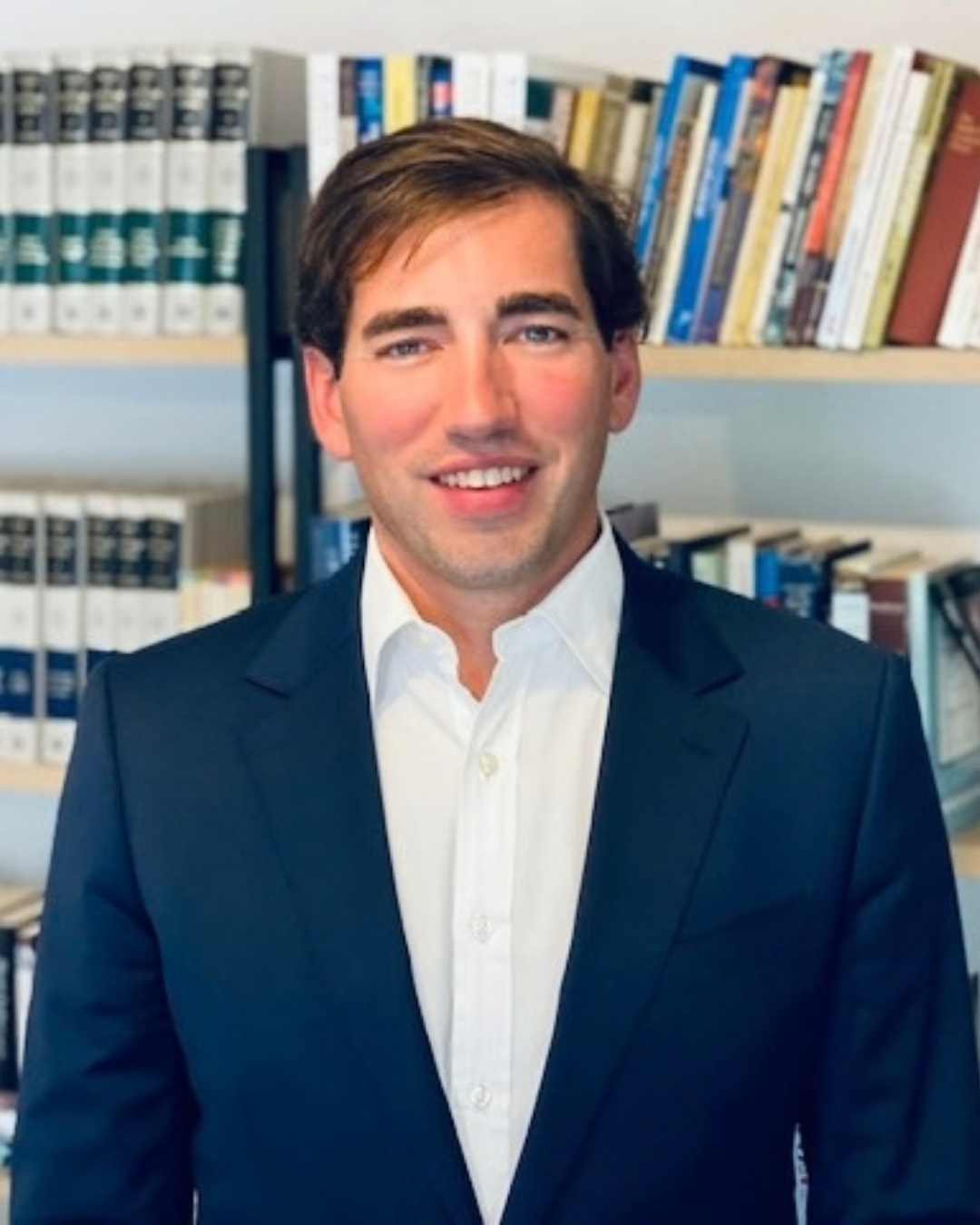 A man in a navy suit and white shirt smiles at the camera in front of a bookshelf.