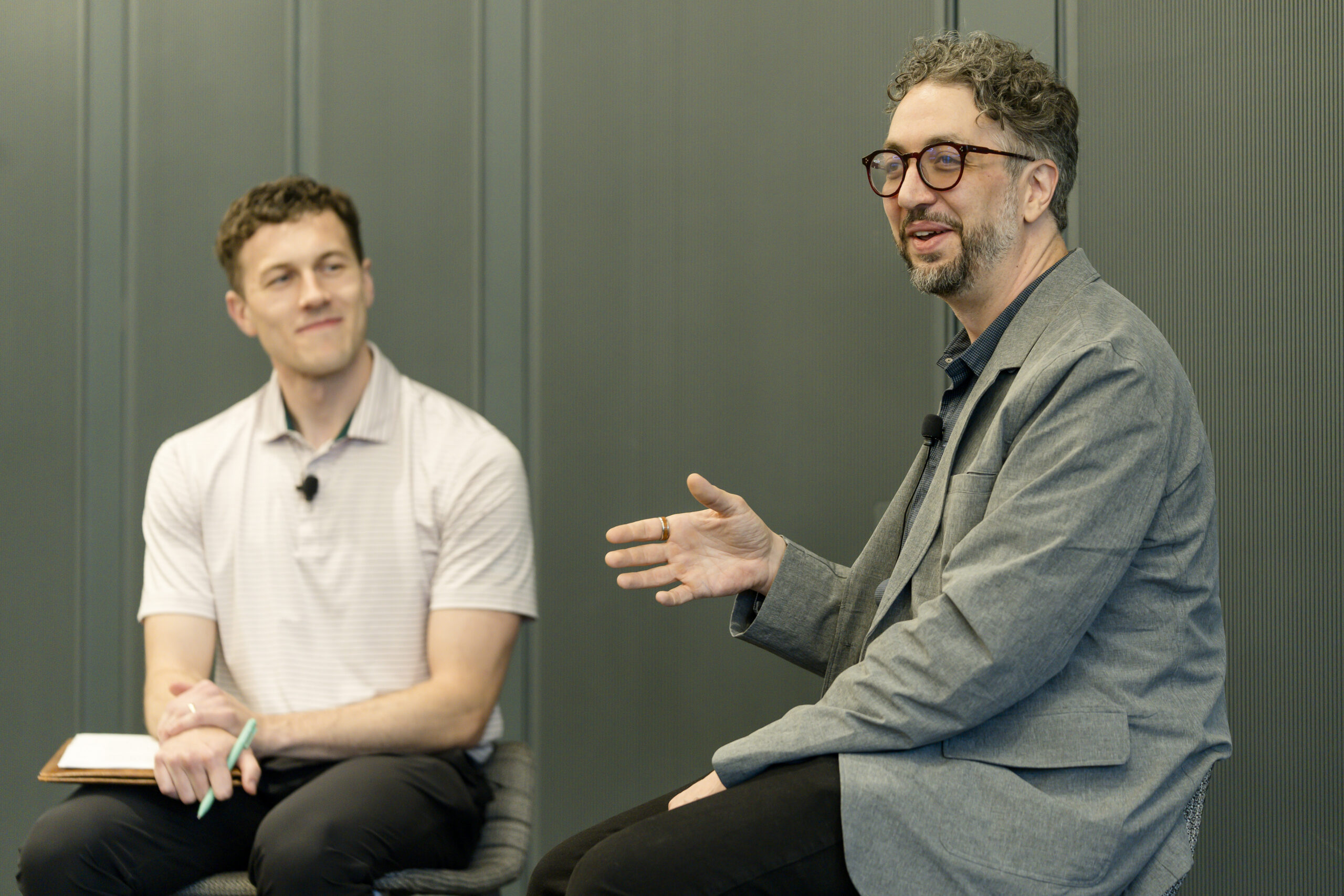 Two people sit in chairs and speak to a room. Both are dressed in business casual attire and have happy expressions on their faces.